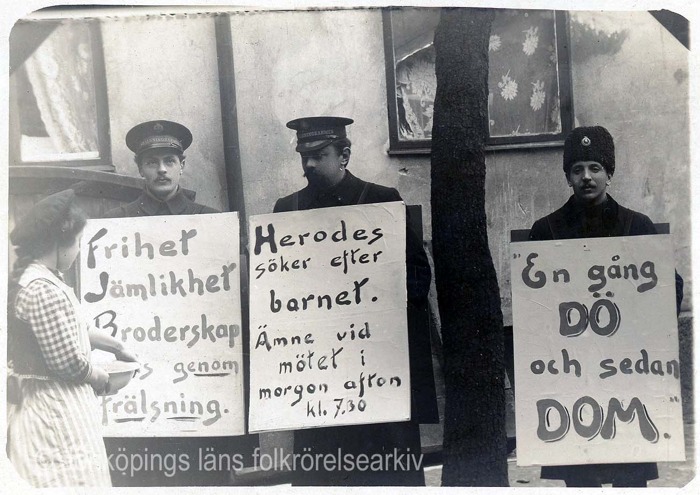 Svartvit foto på tre frälsningssoldater som håller upp skyltar. En kvinna står och läser på en av skyltarna. Tillhör: Frälsningsarmén i Jönköpings arkiv. Svartvit foto på tre frälsningssoldater som håller upp skyltar. En kvinna står och läser på en av skyltarna. Tillhör: Frälsningsarmén i Jönköpings arkiv.