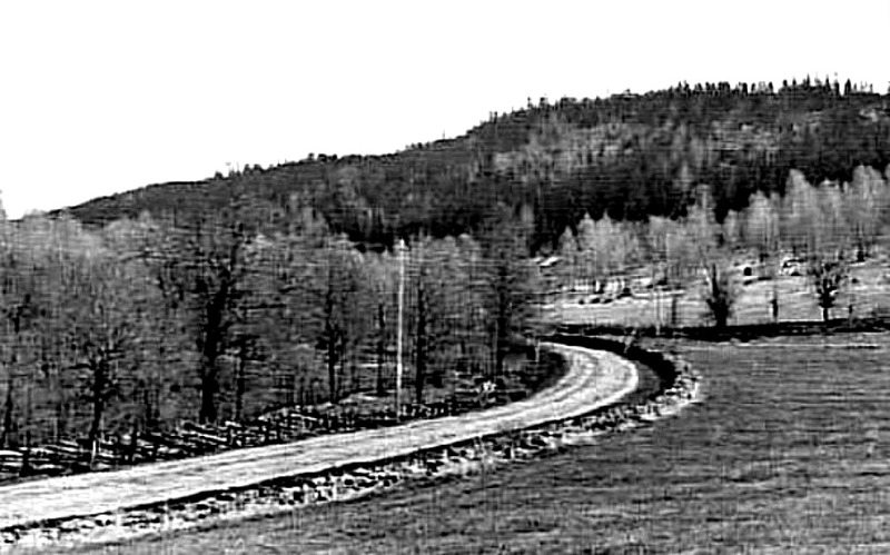 B. 7 (6) Endast digitl bild! Vägen vid hemmanet Sten och nära Fermabron 1930-tal. En bild från 1930-talet av den "nya vägen" som 1877 års väg mot Skövde i dalen mellan bergen kallades länge, innan riksväg 49 byggdes under 1950-talet. Man ser avfarten för fägatan in mot Stenslund till höger med stenmurar på vardera sidan.  Läget för vägen var då ett rejält stycke mer söderut än där riksvägen sedan kom att byggas. Bäcken flyttades mot norr och en ny korsning med både Ljungstorpsväg och dess fortsättning upp mot Stenslund skapades, vilket ödelade den gamla vackra stenbron och förändrade miljön påtagligt. Riksvägens läge kom att hamna i vänster del av bilden ungefär rakt fram från betraktaren räknat.  Hemmanet Sten revs 1938, då Bäckdalen 2 (Petter Lindqvists hus från 1833) revs och man använde virket från Sten för återuppbyggnad. Margit Linqvist, som idag äger det huset berättar att hennes far köpte det tomma huset i Sten 1938, det var ett timmerhus. Han rev det och transporterade stockarna till skiftet för Margits hus. Det gamla Lindqvistska huset revs också men även där återanvändes stockarna och Margits nuvarande hus blev resultatet. Margit minns detaljer när byggnationen skedde bl a en dörr med lucka som hon minns var ställd upp och ner.  Spismuren i Sten stod kvar som ett skelett tills man sprängde den, kvar är en del stenar som markerar var husgrunden låg. (Berättat för Arne Sträng, 2014-02-19. Insatt av Kent Friman, 2014-02-25. Läs mer på www.ljungstorpshistoria.se!