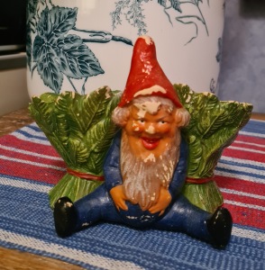 Tomte sittande framför grön kruka. Bredd 15 cm, höjd 11 cm. Färgskav men ändå charmig. 160 SEK