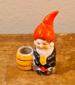 Sittande minitomte vid randig tunna. Lagad luva, annars ok. Höjd 6,5 cm. 30 SEK