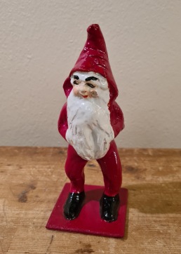 Stående tomte  i röda kläder på platta. Höjd 14 cm. Liten skada på kinden (se bild), annars gott skick. 65 SEK