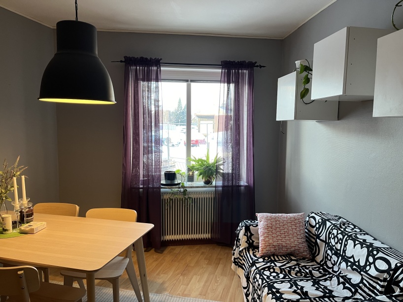 Syntolkning: Ett foto av ett rum med grå väggar, ett fikabord, en liten soffa och ett fönster med lila gardiner.