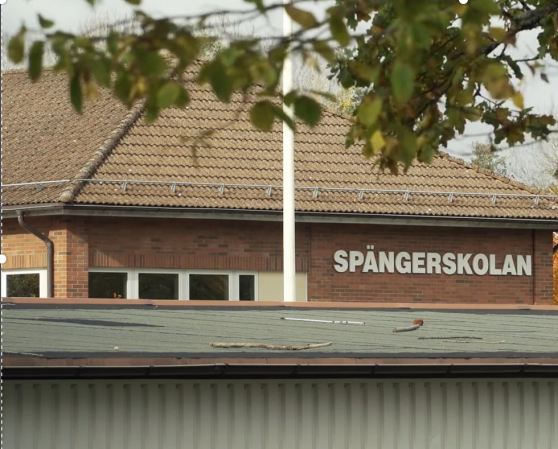 Syntolkning: En tegelbyggnad med texten Spängerskolan. Framför syns en flaggstång och ett hus med plåttak samt en trädgren.