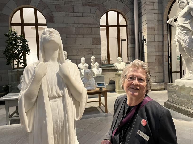 Syntolkning: Ett foto från National museum med en vit staty i naturlig storlek föreställande Fredrika Bremer. Bredvid står en medelålders kvinna i grå kavaj: Jeanette Rangner Jakobsson, intendent på pedagogiska enheten. I bakgrunden syns flera mindre skulpturer.