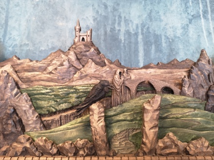 En svart korp i förgrunden till det avlägsna slottet på höjden. Däremellan slingrar sig vägen över djupa ravinen och den trasiga stenbron