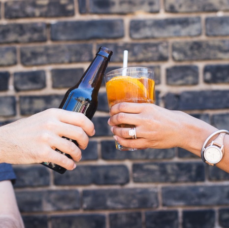 Bild på två händer som skålar, den ena håller i en ölflaska och den andra i ett glas med en drink. Snygg, murad vägg i bakgrunden i rustik stil.