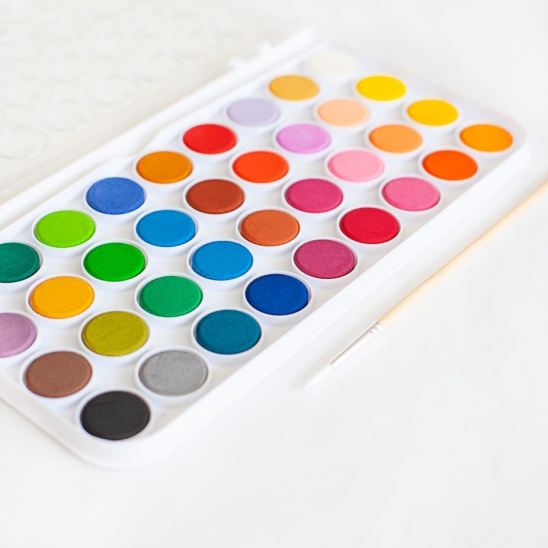 Bild på en oanvänd målarpalett med färgglada vattenfärger.  Paletten ligger på ett vitt bord med en oanvänd pensel bredvid.