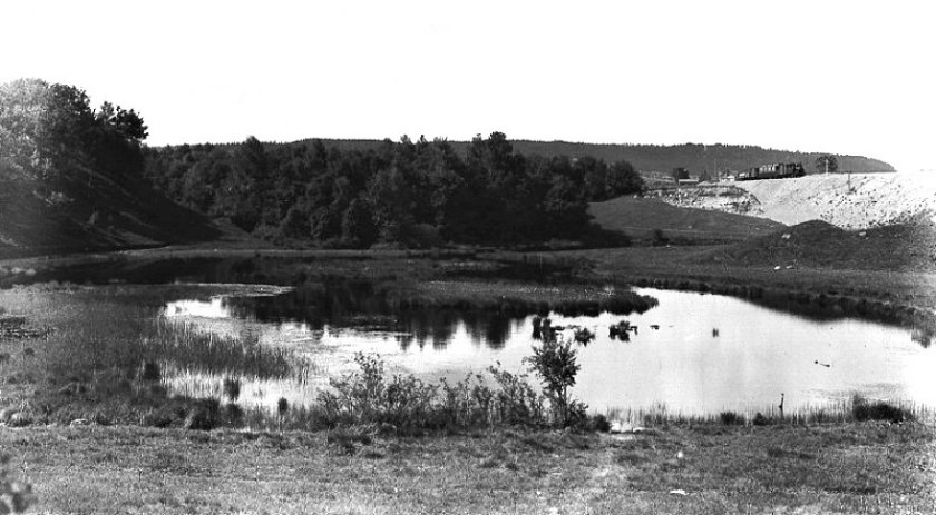 Harkessjön, sedan långt tillbaka igenfylld, låg alldeles nära SAJ-banan och en bit från Varnhemsvägens överfart. Banvaktsstugan skymtar bortom Skövde-tåget som tuffar fram 1905. (Skövde Stadsmuseum)