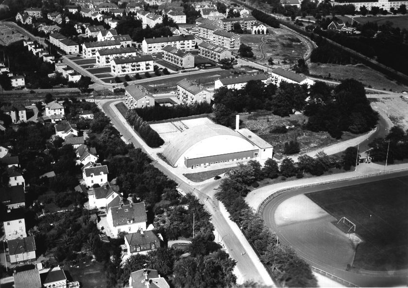 1950-talet efter nyss uppförd idrottshall. SAJ svänger ut från stambanan alldeles efter Vasaplan och passerar Skaraborgsgatan. (Skövde Stadsmuseum)